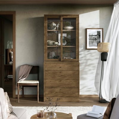 Cosy room with TONSTAD storage. Wooden cabinet, large, with glass doors & drawers. Modern, brown & beige tones. Floor lamp right, chair left.