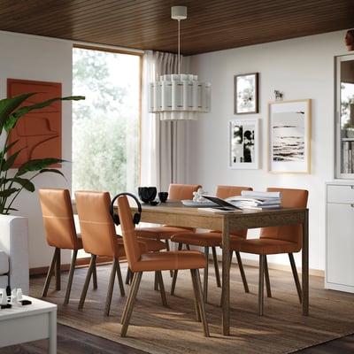 Modern dining room with oak table, six chairs, pendant light, art wall, large window, and plant.