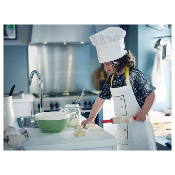 Child wearing white chef hat and apron, rolling out dough on a kitchen counter with bowls and jars nearby.