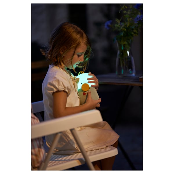 Young girl seated, hugging a blue stuffed toy, wearing a white shirt, in a dimly lit room.