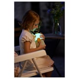 Young girl seated, hugging a blue stuffed toy, wearing a white shirt, in a dimly lit room.