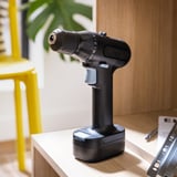 Black ikea TRIXIG cordless drill stands on table, smaller than yellow stool, has rubber grip & two batteries.