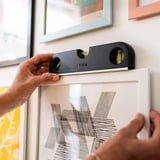 Hands use magnetic spirit level to ensure straight placement of framed art on wall.