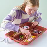 A girl organizes craft supplies in a red TROFAST storage tray with compartments on a table.