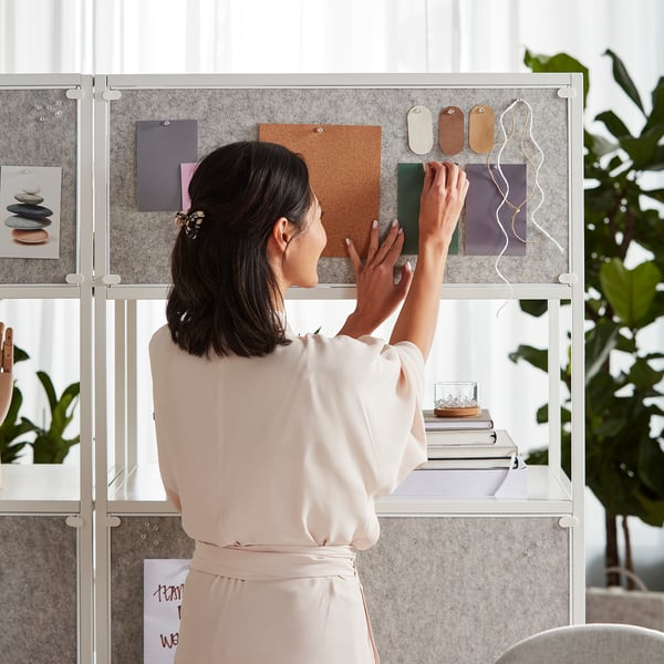 A woman in a beige outfit is hanging a green notepad on the TROTTEN double-sided cabinet. The cabinet shows notice board functionality.