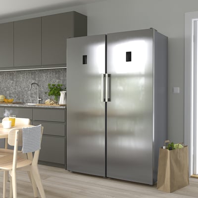 Large modern kitchen with TYNNERÅS stainless-steel refrigerator. Tall, sleek design, next to wooden chairs and grey cabinets.