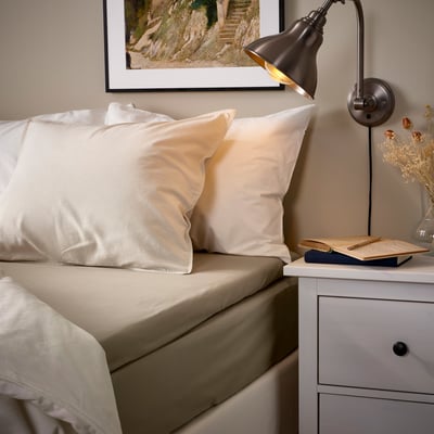 Cozy bed with ULLVIDE beige sheets, white pillows, wall lamp, framed picture, and small nightstand with a book and vase.