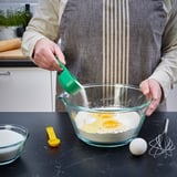 Person using green UPPFYLLD measuring spoon to add ingredient to mixing bowl. Apron worn, other ingredients nearby.