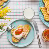 Heart-shaped waffles with sugar, cream, jam on turquoise plate with utensils and flowers.