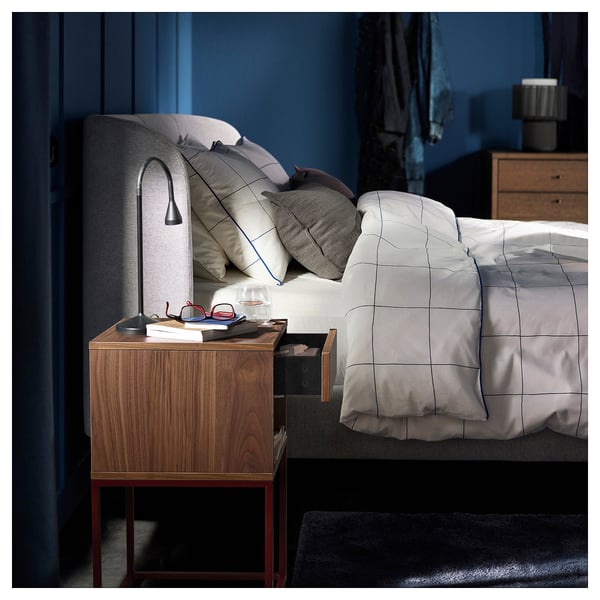 Cozy bedroom with dark blue walls, light gray bedding, and a wooden nightstand with a lamp and book.