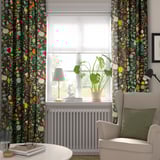 Bright floral curtains behind large window, light gray armchair with green pillow, plant on windowsill with lamp and books.