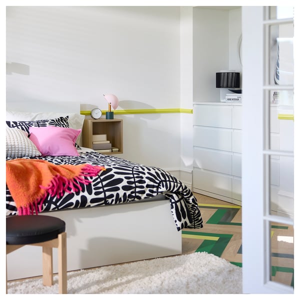 Bright bedroom with white bed, colorful pillows, patterned bedding, wooden stool, nightstand, and a fluffy white rug on wooden floors.