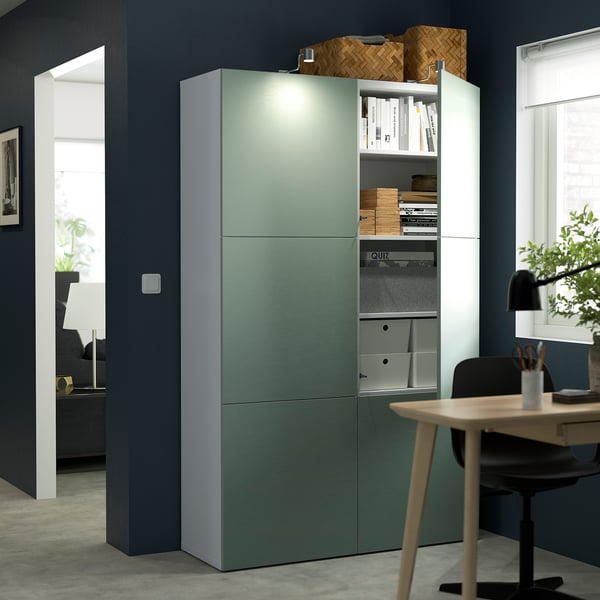 Moderni toimisto, jossa on vihreä BESTÅ-säilytyskaluste, säädettävät hyllyt ja integroidut valot. Mahtuu kirjoja ja tarvikkeita yhdistettynä puiseen työpöytään ja tuoliin.
