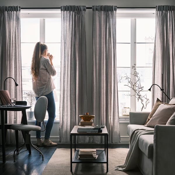 Nainen ikkunan ääressä, jossa on läpikuultavat verhot, lämmin päivänvalo, sohvapöytä kirjojen kanssa.