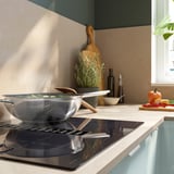 Moderni keittiö, jossa on ruostumattomasta teräksestä valmistettu induktioliesi, vihreitä vihanneksia, puisia astioita, pulloja ja tuoretuotteita.