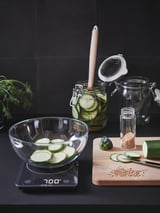Keittiön kokoonpano: asteikko 700 g, viipaloidut kurkut, siemenet, veitsi, suolakurkku, mausteet, yrtit, tumma tausta.