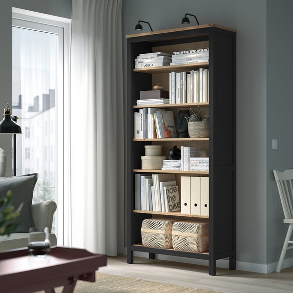 Musta HEMNES-kirjahylly ikkunan lähellä, jossa on kirjoja ja koristeita, korkea, tuoli- ja pöytäpöytä.