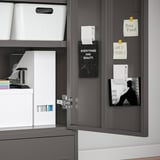 Avoin harmaa kaappi, jossa on järjestettyä säilytystilaa: valkoisia roskakoreja, muistiinpanoja ja motivaatiojulisteita.