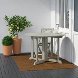 Moderni patio, jossa pyöreä LYDERSHOLM-pöytä, tuolit, kasvit ja sitruunapuu harmaan seinän ja valkoisen ikkunan vieressä.