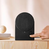 Henkilö painaa nappia mustassa, kaarevassa bluetooth-kaiuttimessa, joka istuu puupinnalla, mikä esittelee sen intuitiivista ohjaustoimintoa.