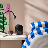 Musta bluetooth-kaiutin kevyellä pöydällä, värikäs taide lähellä, eloisat vuodevaatteet.