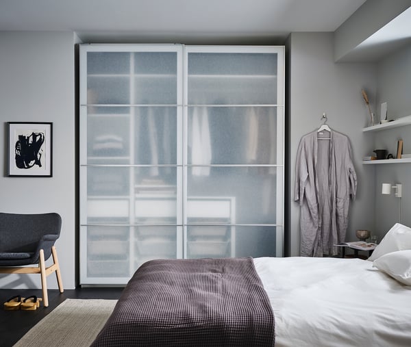 Moderni harmaa makuuhuone, jossa on iso PAX-vaatekaappi, lasiliukuovet, riippuva kylpytakki, harmaa sänky valkoisilla liinavaatteilla, nojatuoli ja seinätaide.