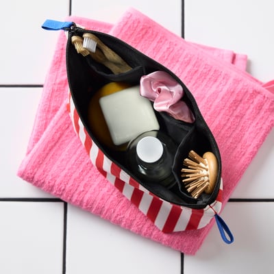 Punainen/valkoinen raidallinen toilettilaukku sinisellä vetoketjulla vaaleanpunaisella pyyhkeellä.