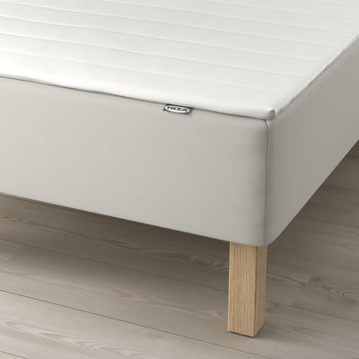 Valkoinen sängynpohja puisilla jaloilla, IKEA-etiketti näkyvissä sivulla.