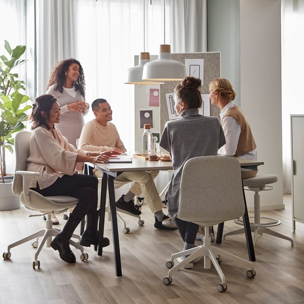 Viisi ihmistä hymyilee beigen TROTTEN-työpöydän ympärillä, mikä on tehokas ja tilaa säästävä kotiin tai toimistoon.