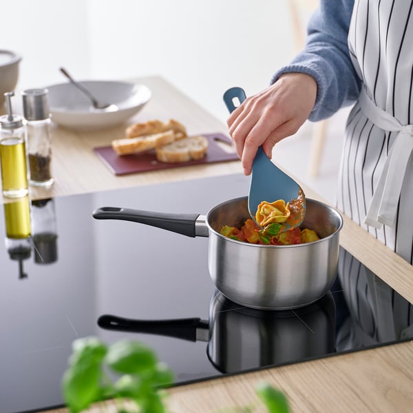 Henkilö, joka keittää pastaa kattilassa sinisellä lastalla. Puinen työtaso, jossa leipää, öljyä ja yrttejä taustalla.