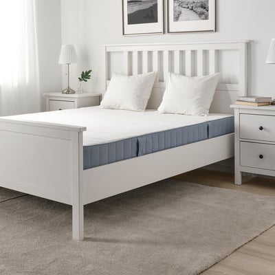 Minimalistinen makuuhuone, jossa valkoinen VESTMARKA-patja, kaksi tyynyä, yöpöytä lampuilla, kirjoja ja kasveja, matto.