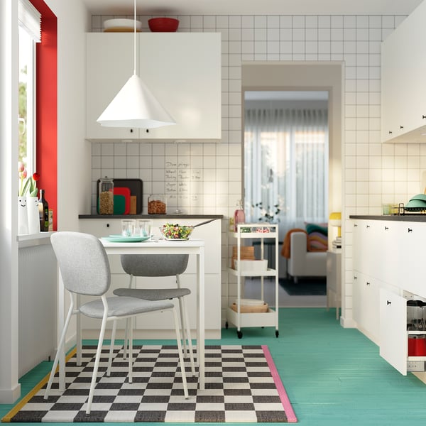 Moderni keittiö, jossa valkoinen VIHALS-pöytä ja kaksi harmaata tuolia mustavalkoisella matolla. Kulhollinen salaattia istuu pöydällä punaisella koristellun ikkunan vieressä.