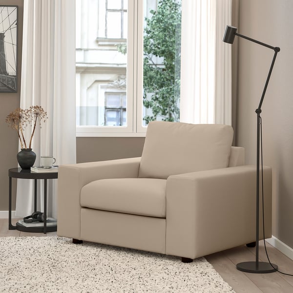 Moderni beige sohvatuoli, minimalistinen muotoilu, suuri ikkunatausta, korkea musta lamppu, sivupöytä kuivatuilla kukilla, viihtyisä kotiympäristö.