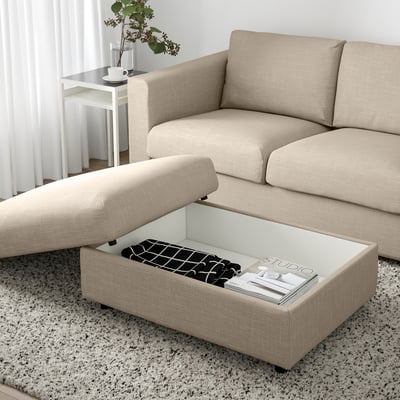 Beige VIMLE-sohva, jossa on ottomaani. Ottomaanien istuin on nostettu, mikä paljastaa säilytystilan, jossa on lehtiä ja taitettua kangasta.