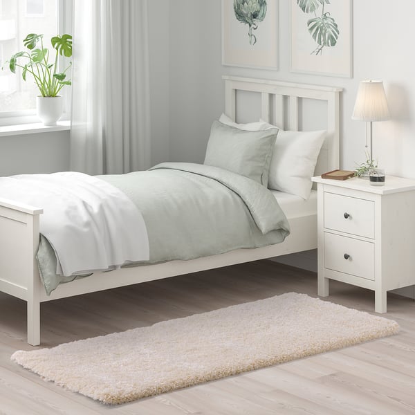 Makuuhuoneessa valkoinen sänky, muhkea VOLLERSLEV-matto, vihreät liinavaatteet, kasvi ja lamppu.