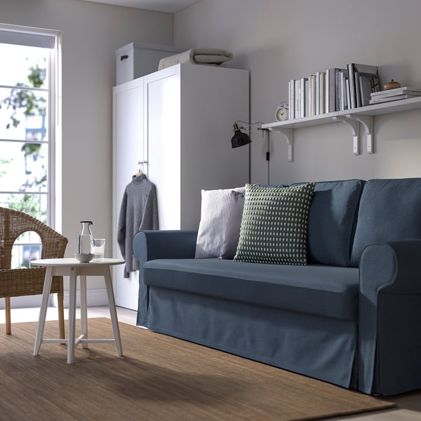 Moderni olohuone, jossa VRETSTORP-sininen vuodesohva, tyynyt, sivupöytä, korituoli, kirjahylly ja kello. Minimalistinen, mukava.