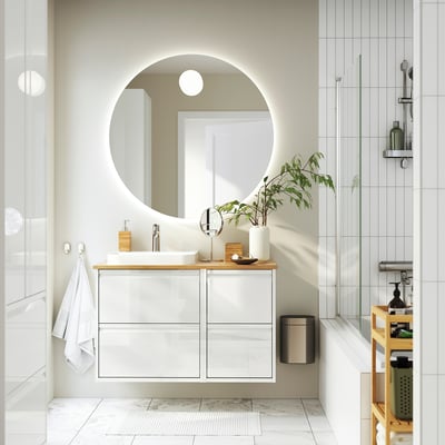 Modernt badrum med det vita skåpet ÄNGSJÖN, träbänk, rund spegel, växt och dusch i glas.