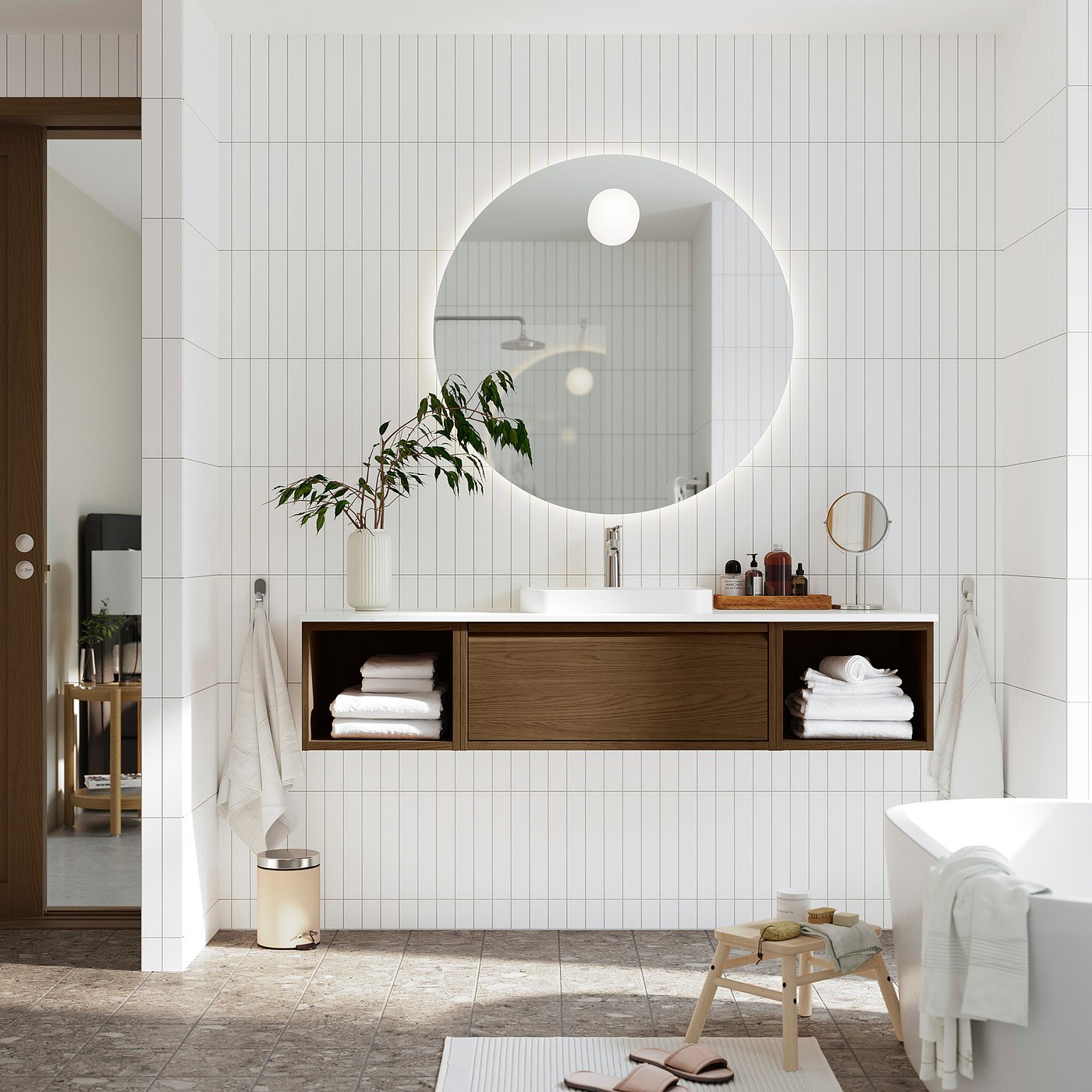 Modernt badrum med svävande skåp i brun ek, vit bänk, rund spegel, vita handdukar, grön växt och badkar.