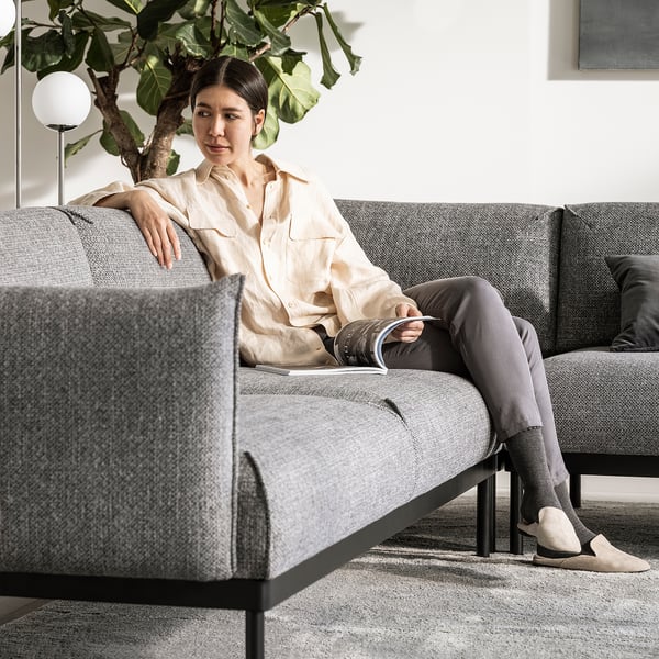 Kvinnan kopplar av på modern grå ÄPPLARYD soffa, ljus skjorta, mörka byxor.