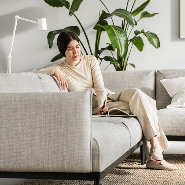 Bekväm kvinna som skriver på soffan, grädde blus och byxor, stor bladväxt bakom, vit lampa, modern grå soffa, mysig atmosfär.