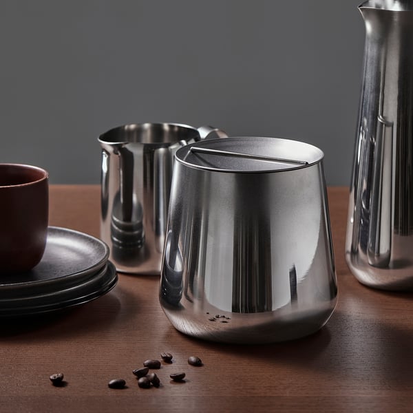 Flera silverburkar och en kaffekopp på ett mörkt bord med utspridda bönor. Den polerade AFTONHAJ-burken reflekterar.