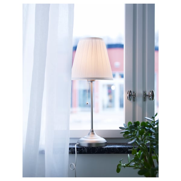 En lampa med en beige nyans på en fönsterbräda, diffust ljus, vita gardiner och en grön växt.