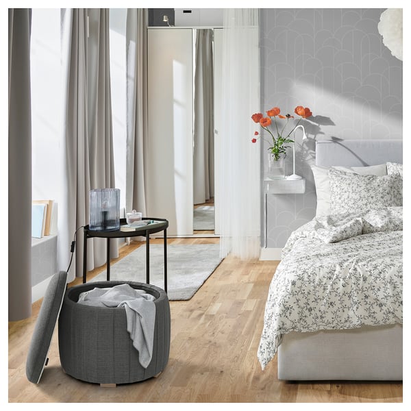 Modernt sovrum med trägolv, stort fönster, blommig säng, grå ottoman, runt bord med röda blommor, vit matta och nattduksbord.