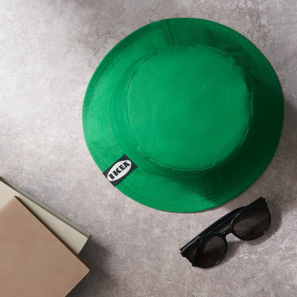 En knallgrön hatt med IKEA-etikett ligger på en grå yta, tillsammans med en bärbar dator och solglasögon.