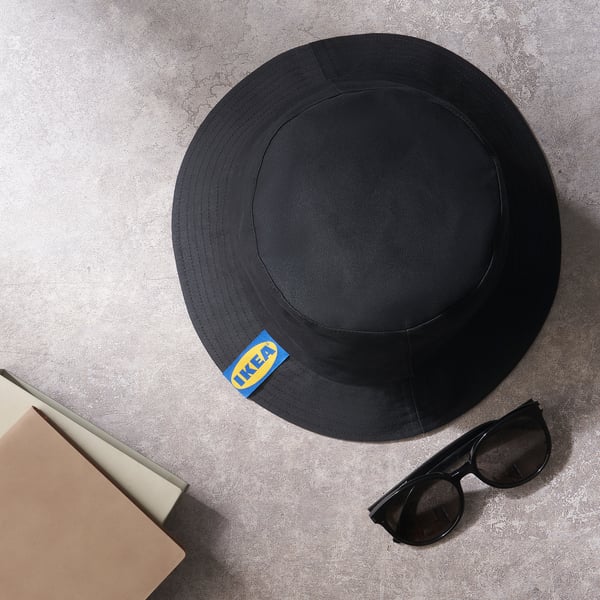 Svart hatt med IKEA-logga, solglasögon och öppen bok på grå yta.