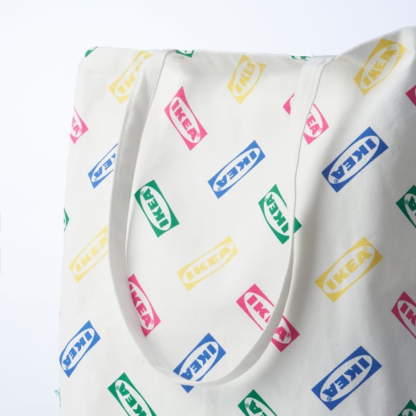 Vit återanvändbar tygkasse i bomull med färgglada IKEA-logotyper utspridda mönster, med två robusta handtag, perfekt för daglig användning.