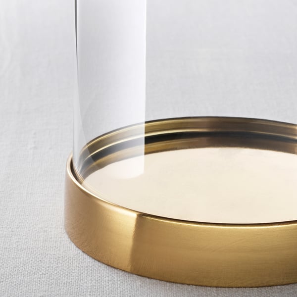 Närbild av en elegant, klar glaskupol med en gyllene metallisk bas, som reflekterar ljus och visar upp ett dekorativt föremål inuti.