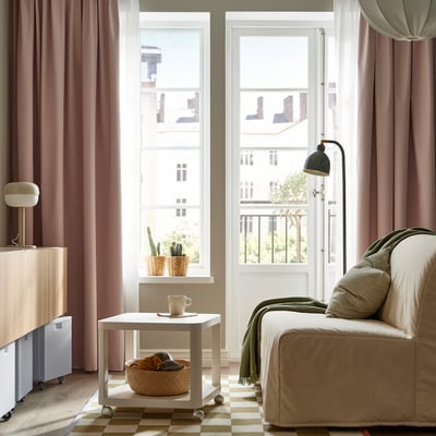 Stora, mjuka rosa BENGTA-gardiner ramar in ett rum med en bekväm soffa under ett högt fönster och erbjuder en mysig och väl upplyst atmosfär.