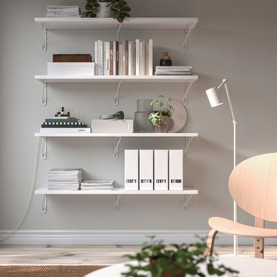 En modern studiehörna med vita BERGSHULT-hyllor med böcker, inredning och växter. En stol och golvlampa i trä fulländar den minimalistiska designen.