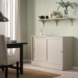 BERGSHULT hylla i beige rymmer tekanna, vaser, växter; ovanför beige skåp, nära svart bord.